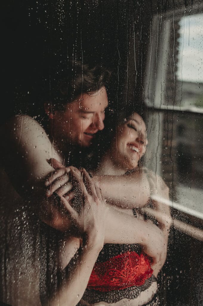 Playful and steamy moment between a couple during their couples boudoir session at Helios Boudoir Photography, smiling and embracing behind a rain-covered shower window.