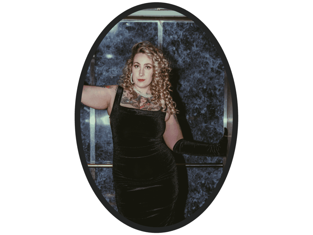 Portrait of a woman with long curly blonde hair wearing a black velvet dress, sheer neckline, and pearl necklace, confidently posing in front of a blue marbled wall inside an elevator, framed in an oval.